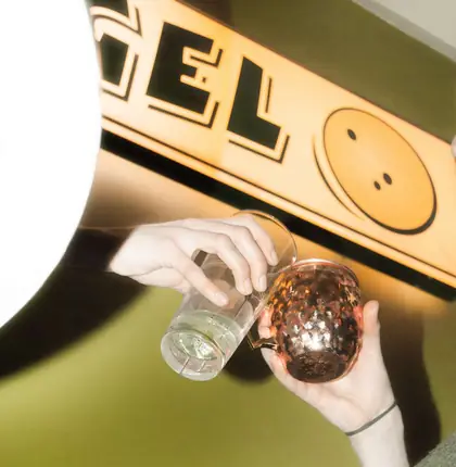 Zwei Hände stoßen mit einem Glas und einem Kupferbecher an, darüber ein warm beleuchtetes Schild an olivgrüner Wand.